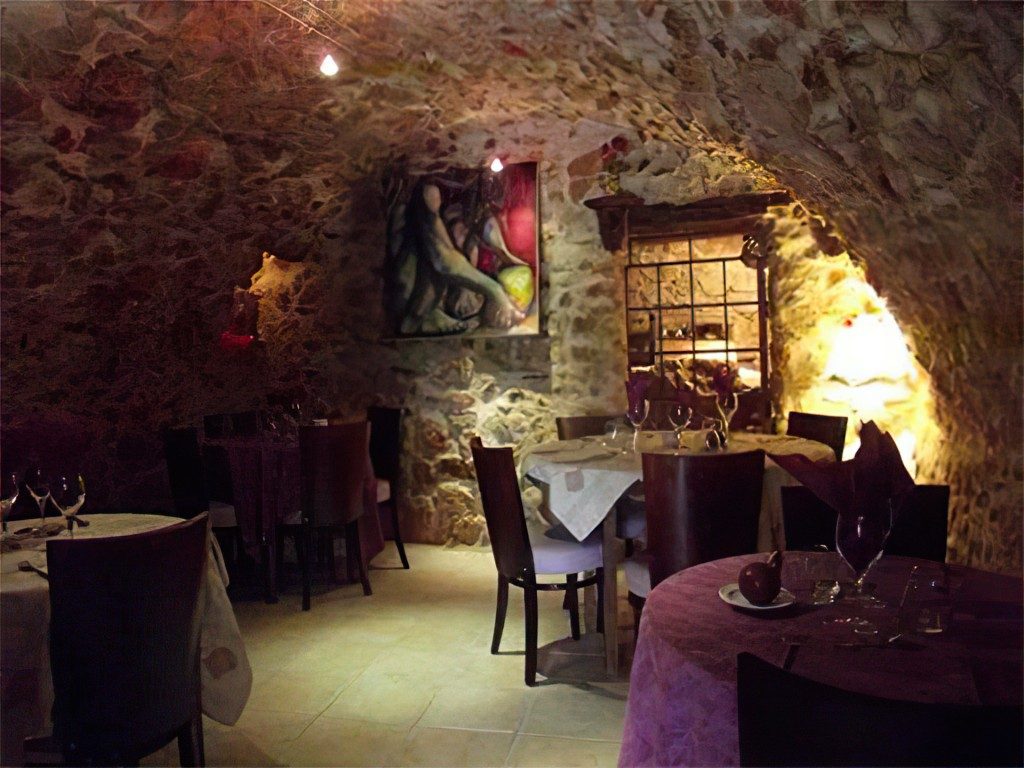 La salle voûtée en pierre : lumière chaude, tables dressées, tableaux aux murs